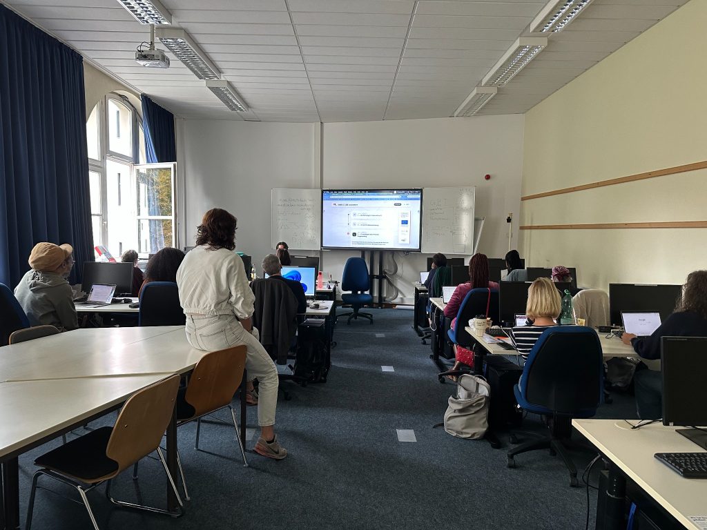 Lerngruppe an Computerarbeitsplätzen mit großem Bildschirm im mediaLab-Kursraum.